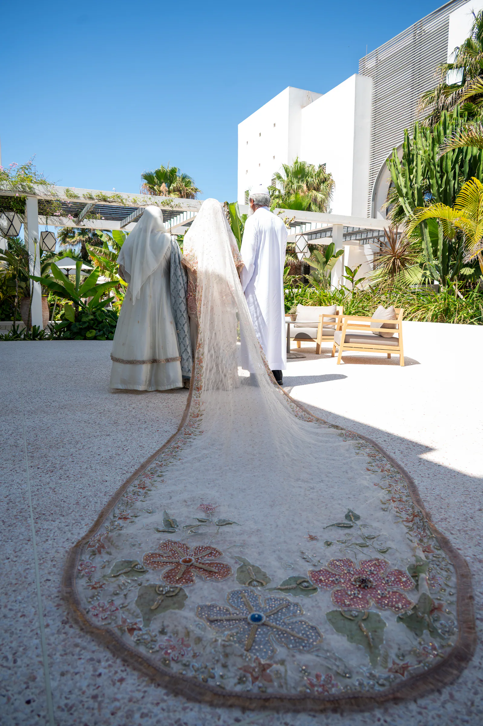 Une mariée avance dans une cour ensoleillée, vue de dos